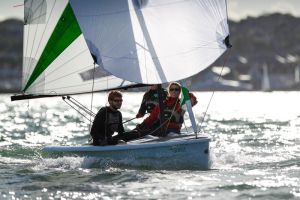 Three people sail a small boat with a white and green sail on a sunny day. The water sparkles around them, and distant boats and shoreline are visible in the background.