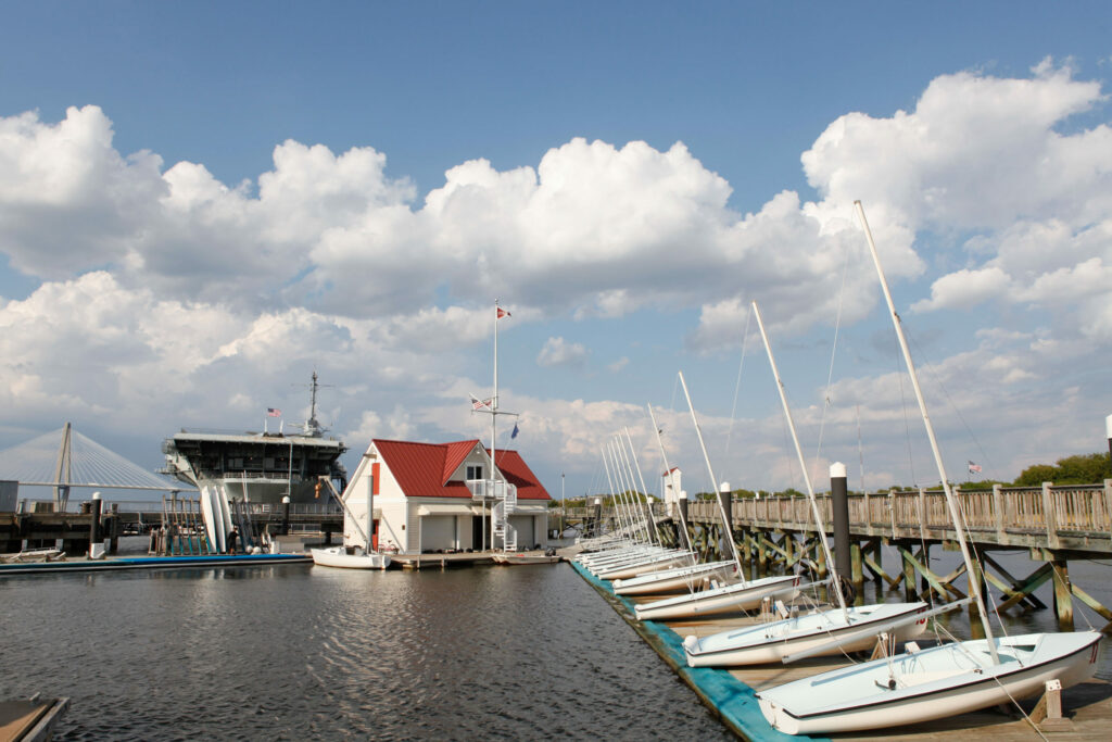 Sailboats are docked along a wooden pier beside a small white building with a red roof, with a larger ship and a bridge in the background under a blue sky with scattered clouds.