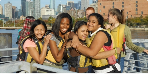 A group of smiling teens wearing yellow life jackets stand on a dock by the water with a city skyline in the background, hugging and enjoying a sunny day outdoors.
