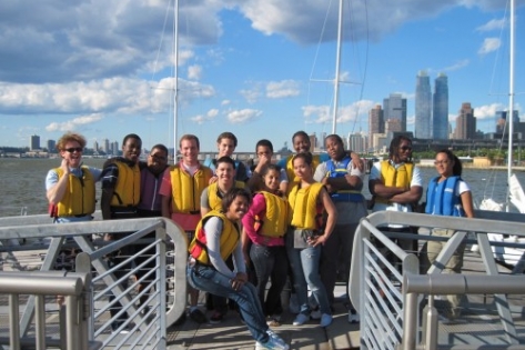 A group of people wearing life jackets pose and smile on a dock by the water, with a city skyline and sailboats in the background under a partly cloudy sky.