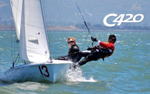 Two sailors wearing gear balance on the edge of a small sailboat marked “13” as it glides on open water; the text “C420” appears in the sky above them.