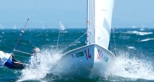 A sailboat with the number 14 on its bow races through choppy blue water, creating a large splash. Two sailors are on board, leaning out to balance the boat. The sky is clear and other boats are visible in the distance.