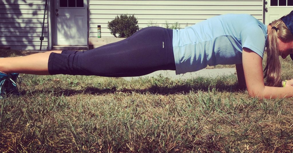 A person wearing a blue shirt and black leggings is doing a plank exercise on grass outside, with a house and door visible in the background.