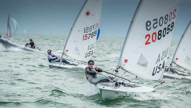 Sailboat racers compete on choppy water, leaning out to balance their small white boats with USA sail numbers visible, under a cloudy sky during a regatta.