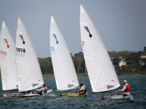 Four small sailboats, each with a single sailor, race side by side on a body of water. The sails are white with colored insignias and numbers. Trees and buildings are visible in the background under a clear sky.