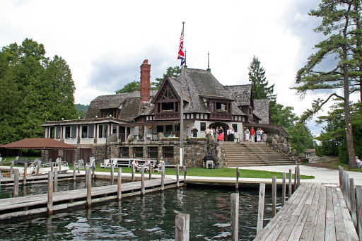 A large, rustic lakeside house with a dock in front, surrounded by trees. People are gathered on the stone steps leading to the entrance. An American flag flies atop the roof under a partly cloudy sky.