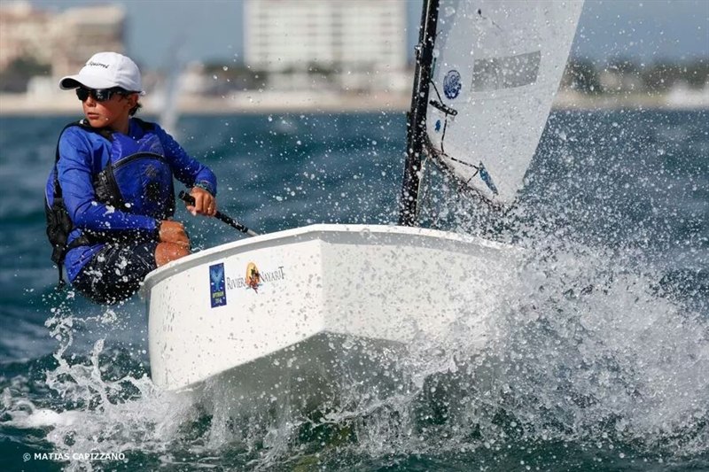 A young sailor wearing sunglasses and a blue shirt navigates a small white sailboat through choppy water, with splashes around the boat and a cityscape blurred in the background.