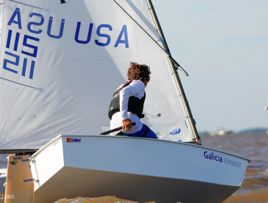 A sailor navigates a small white sailboat labeled 'Galicia EMINENT' in brownish water. The sail displays 'USA' and the number 115211. The person wears a life vest and looks ahead while holding the boat’s tiller.