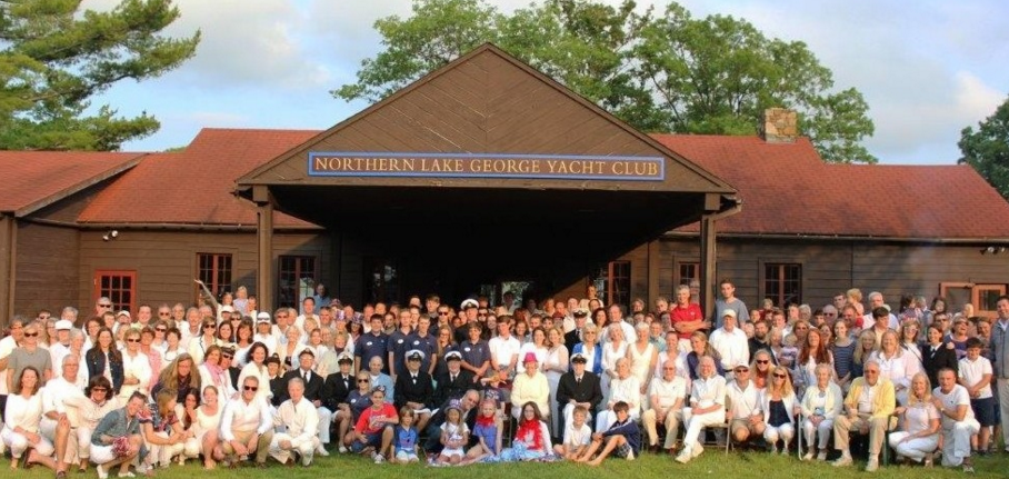 A large group of people gather for a photo in front of a rustic wooden building with a sign reading 'Northern Lake George Yacht Club' on a sunny day.