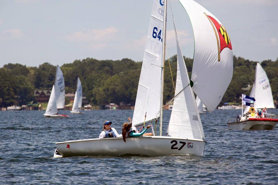 A white sailboat with the number 27, carrying two people in life jackets, sails on a lake with other sailboats nearby. Trees and houses line the distant shore under a partly cloudy sky.