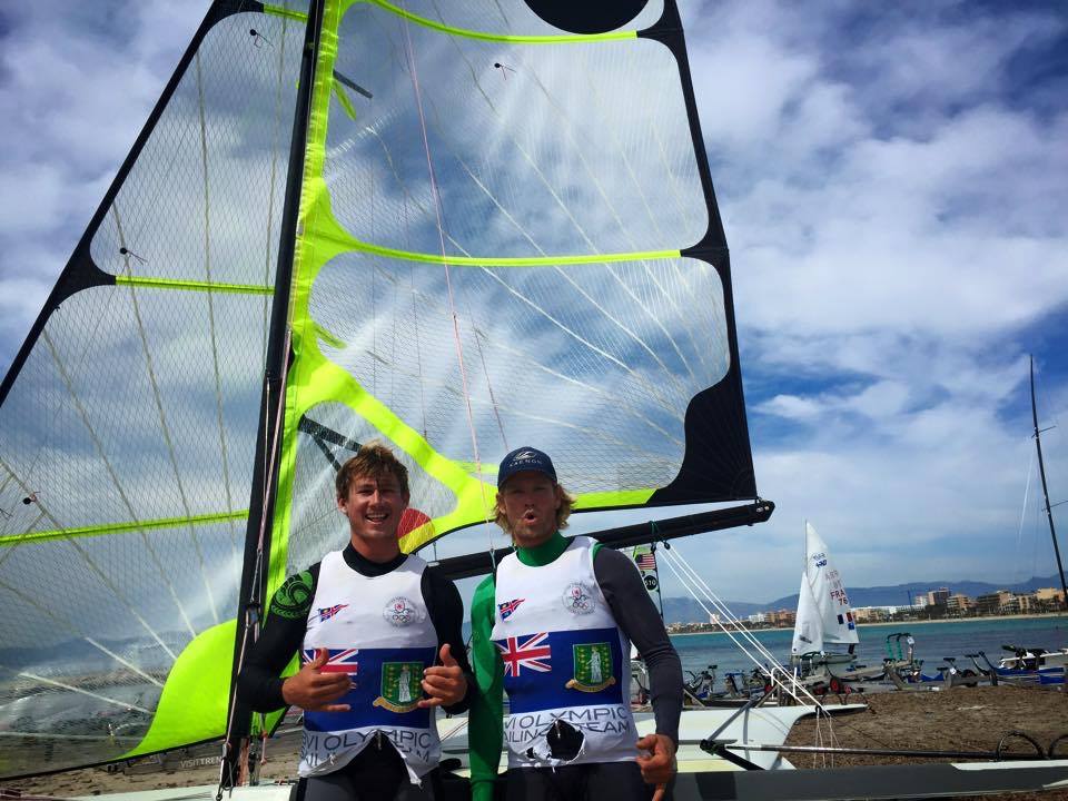 Two sailors in wetsuits and team vests stand smiling in front of a bright green and black sailboat on a beach, with other boats and a cloudy sky in the background.