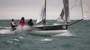 Three people are sailing a small boat named 'Jackpot' with the number 16 on the bow, moving quickly through choppy waters under a cloudy sky.