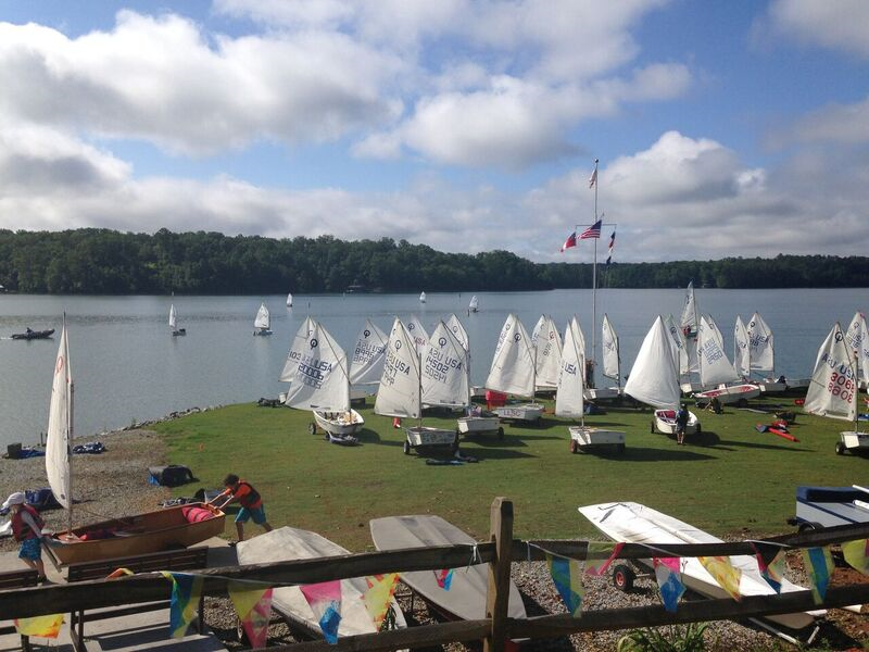 Dozens of small sailboats are parked on a grassy area beside a lake, with more boats sailing on the water. Colorful pennant flags hang from a wooden fence in the foreground under a partly cloudy sky.