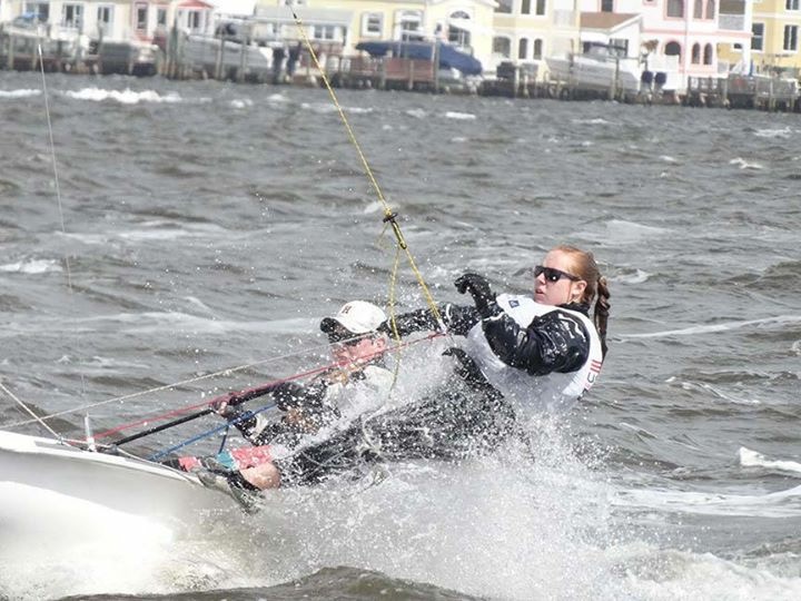 Two people sail a small boat on choppy water, leaning out over the side in wet weather gear. Water splashes around them. Colorful buildings line the shore in the background.