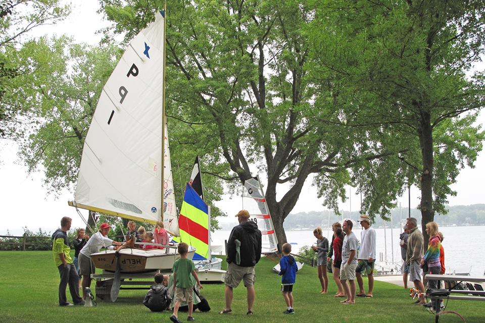 A group of adults and children gather on a grassy lakeshore near two small sailboats. Some people stand while others sit on picnic tables. Trees provide shade, and a colorful flag and calm lake are visible in the background.