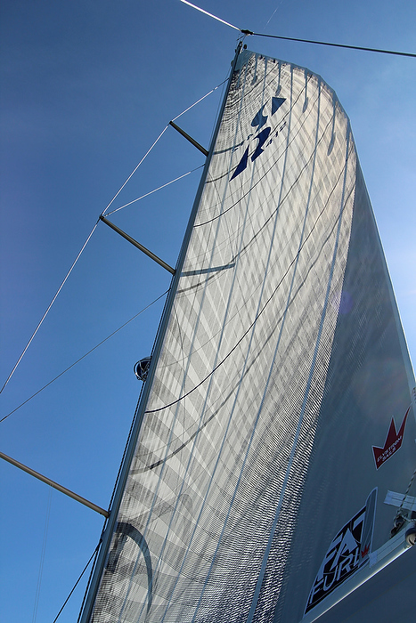 A close-up view looking up at a tall sail on a sailboat against a bright blue sky, showing detailed sail material, rigging lines, and sailboat logos.