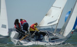 Three people wearing life jackets are actively maneuvering a small sailboat in choppy waters, leaning over the side as the boat tilts, with another sailboat visible nearby under a clear sky.