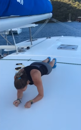 A woman in athletic wear holds a plank position on the deck of a boat, with water and forested hills visible in the background.