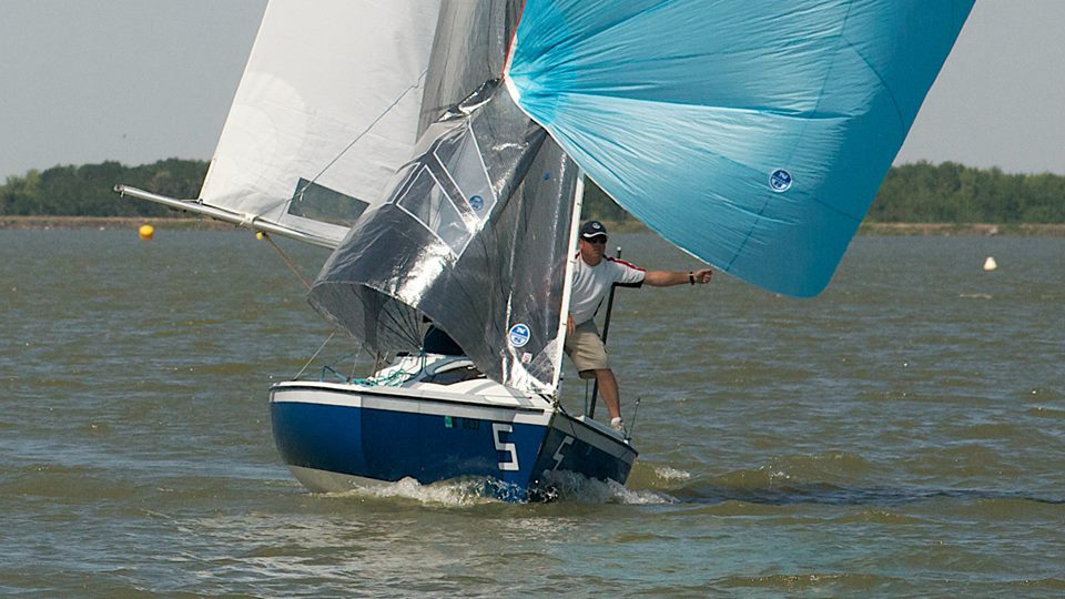 A sailor on a blue and white boat numbered 5 adjusts light blue sails on a windy day, with trees and buoys visible in the background on the water.