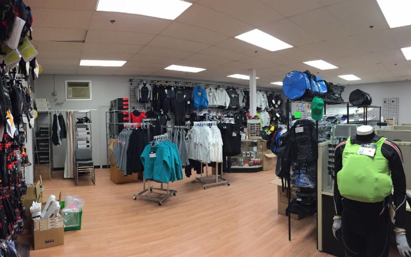 A retail store interior displays various outdoor clothing and gear, including jackets, backpacks, and accessories arranged on racks and shelves. A mannequin wearing a bright green jacket stands in the foreground.