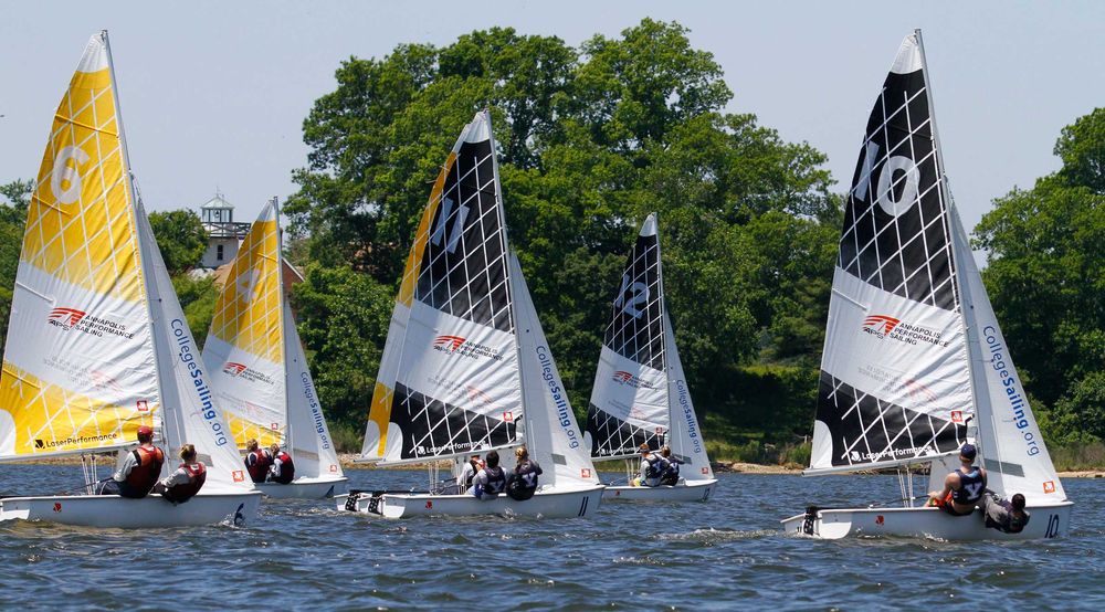 Several small sailboats with colorful, numbered sails compete on a lake. Each boat has one or two sailors. Green trees and a building with a lighthouse are visible in the background under a clear sky.