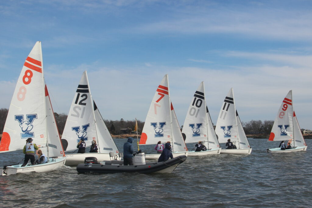 Several small sailboats with numbered sails and 'V' logos, each carrying two sailors, compete on a lake. A motorboat with people in life vests is nearby. The sky is partly cloudy and trees line the shore.
