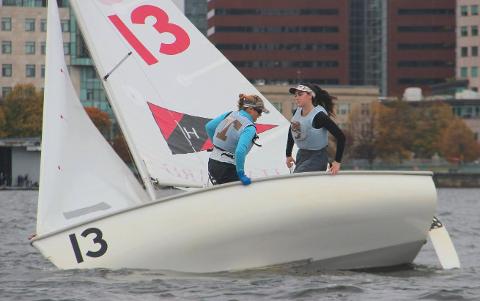 Two sailors in life vests are racing a small sailboat with the number 13 on the hull and sail, leaning as the boat tilts on the water, with city buildings in the background.