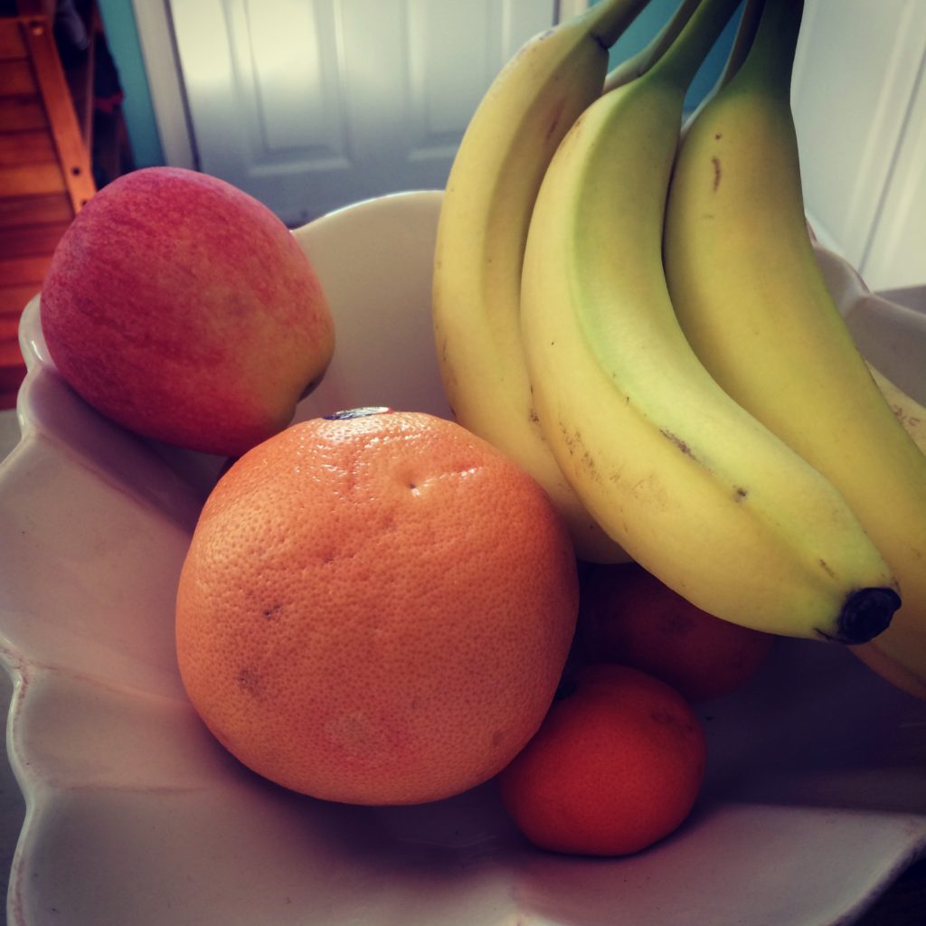 A white bowl holding a bunch of bananas, a grapefruit, an apple, and two small oranges, sitting on a table indoors with soft natural light.