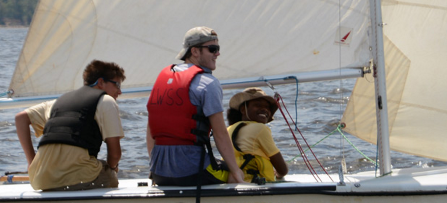 Three people wearing life jackets are sailing a small boat on a sunny day. One person is steering, another handles the sail, and the third smiles at the camera. The water and blurred shoreline are visible in the background.