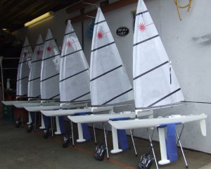 Six model sailboats with white sails and black stripes are lined up indoors on stands, each with a remote control placed underneath. The boats are arranged side by side in a workshop or garage.