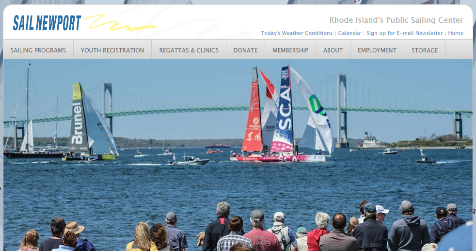 A crowd watches sailboats racing on the water with a large bridge in the background, under a clear sky, at a public sailing event hosted by Sail Newport in Rhode Island.