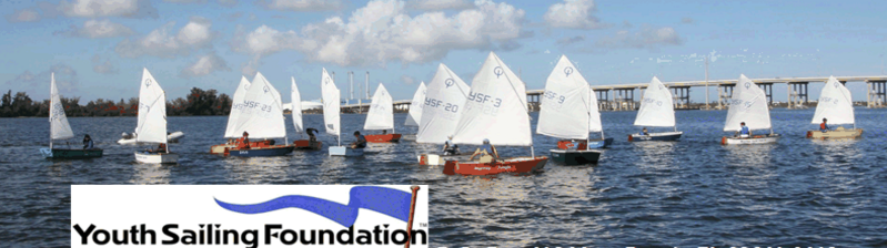 A group of young sailors in small sailboats with white sails navigate on a calm body of water under a partly cloudy sky. A bridge and trees are visible in the background. A Youth Sailing Foundation logo appears in the corner.