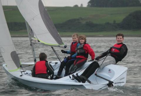 Four children in red and black life jackets are sailing a small white sailboat on a lake, with green hills and trees visible in the background. Two of the children are sitting on the edge, leaning out over the water.