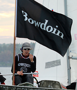 A person wearing sunglasses and a 'Bowdoin' jersey sits on a sailboat, holding a whistle to their mouth. A large black flag with 'Bowdoin' in white lettering waves behind them.