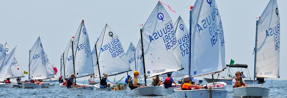 A group of children sail small boats with white sails marked 'USA' on a calm body of water during a sailing race, under a clear sky.