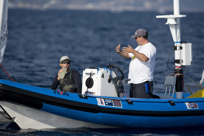 Two people are on a blue motorboat on the water; one is sitting and drinking from a bottle, the other stands and gestures with both hands. The boat has a “GBR” sign, and the weather appears calm and sunny.