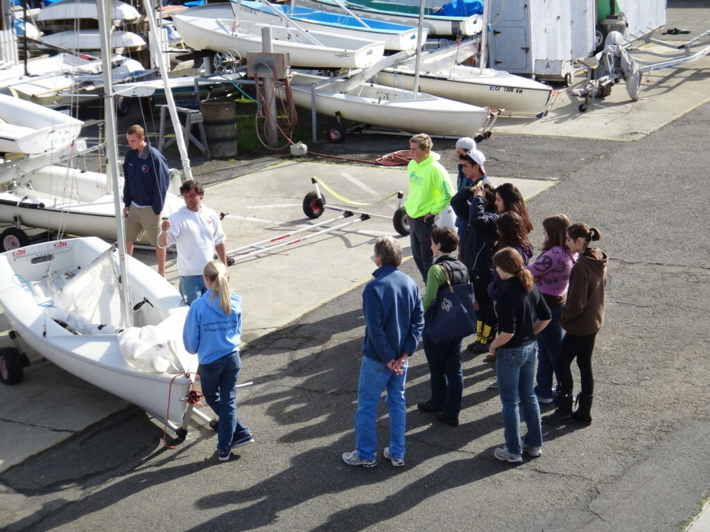 A group of people stand in a semicircle around two instructors next to a small sailboat in a boatyard, listening attentively. Several other boats and equipment are visible in the background.