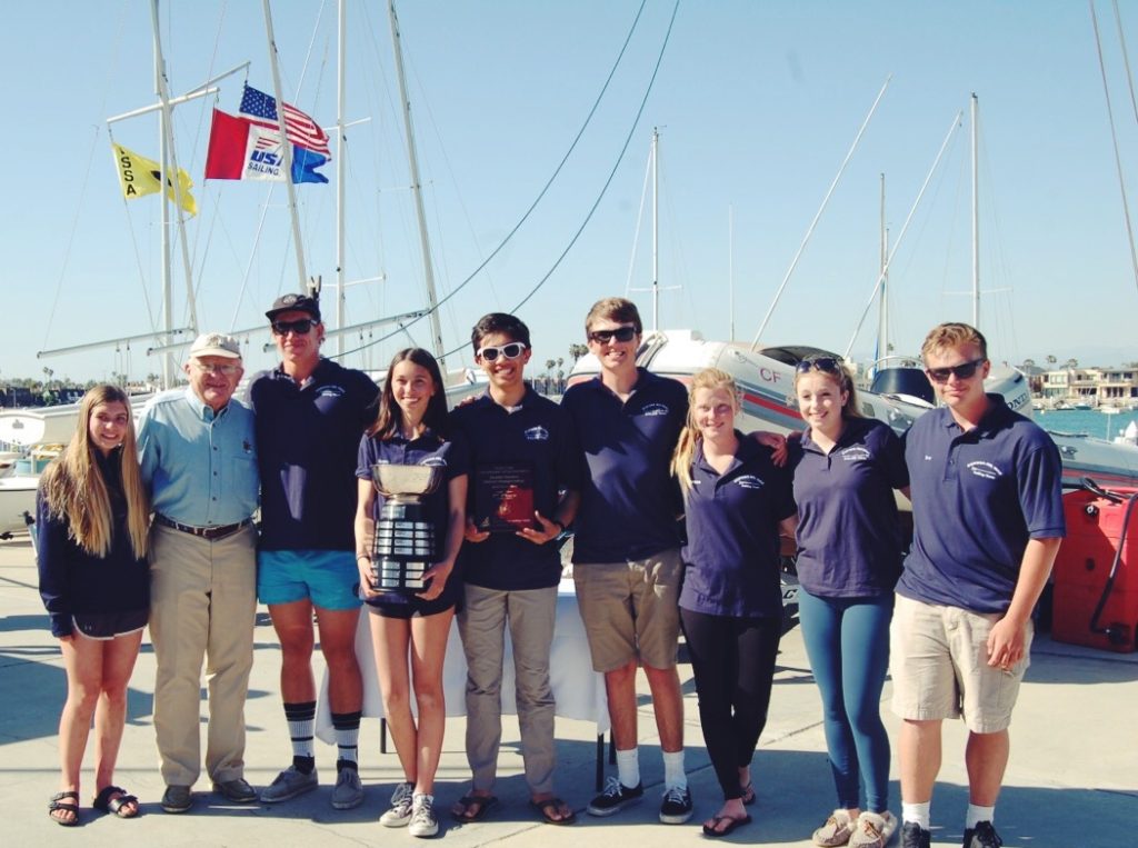 A group of eight people, some holding trophies, stand together smiling in front of sailboats and flags at a marina on a sunny day. All but one are wearing matching navy shirts and shorts.