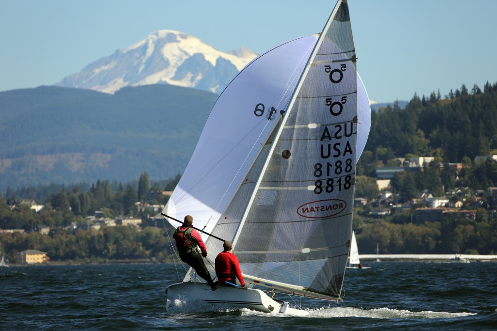 Two people sail a small boat with a white spinnaker and “USA 8818” on the sail across a lake, with a snow-capped mountain and a tree-lined shoreline in the background under a clear sky.