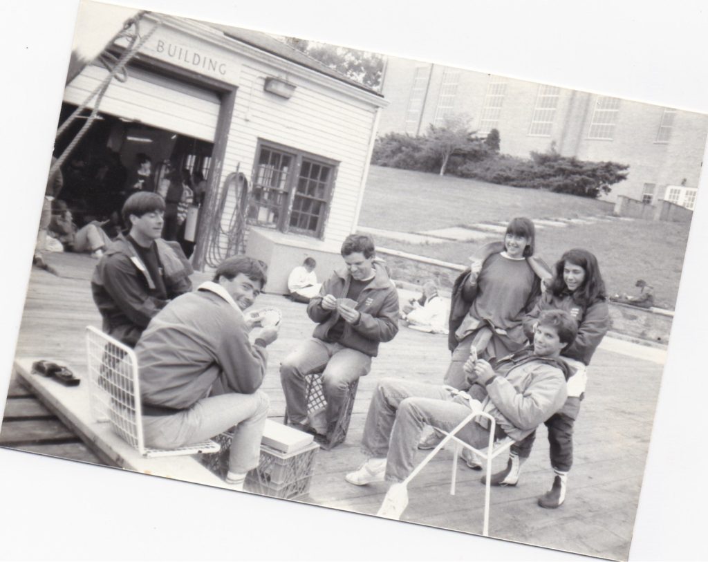 A black-and-white photo of a group of young people sitting and standing outside a building. Some are seated on crates, playing cards, while others stand behind them, smiling. A grassy slope and more people are in the background.
