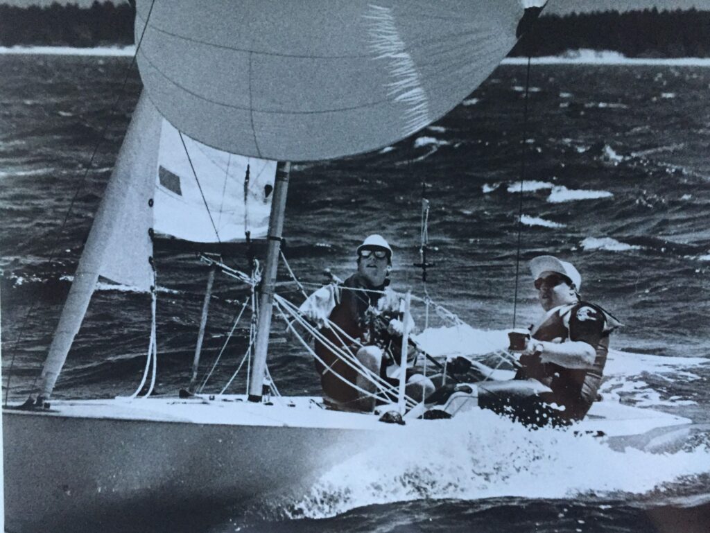 Black and white photo of two people sailing a small boat on choppy water, wearing life jackets and sunglasses, with the sail billowing and water splashing around the boat. Trees line the distant shore.