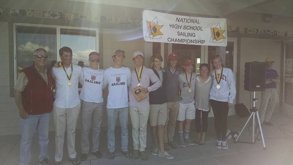 A group of nine high school students wearing medals stand in front of a 'National High School Sailing Championship' banner, smiling and posing for a photo. An adult stands beside them, and one holds a plaque.