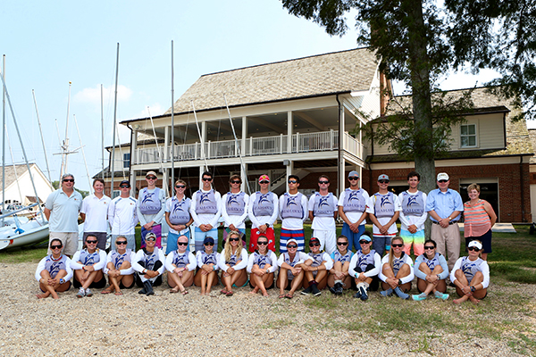 A large group of people pose together outdoors in front of a two-story building, with some standing and others sitting. Many wear matching purple shirts and sunglasses, suggesting they are part of a team or group event.