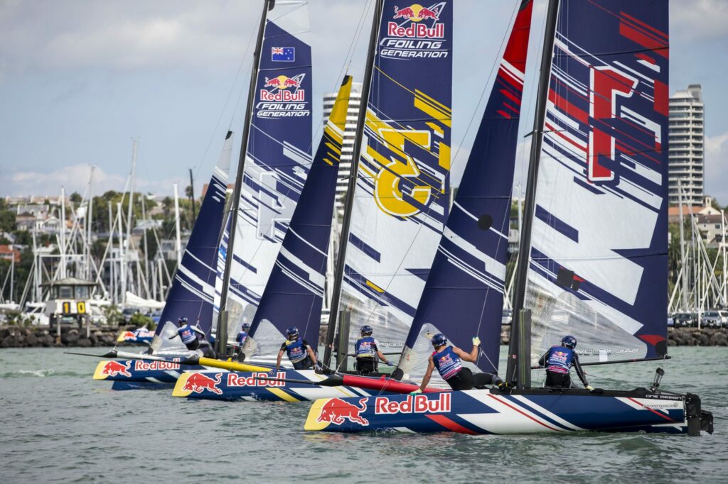 Four Red Bull-branded sailboats with colorful sails race side by side on the water; sailors are focused, and a marina with docked boats and buildings is visible in the background.
