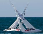 Two sailboats with white sails appear to be crossing very closely on the ocean, their sails tilted and almost touching, creating an X shape against a blue sea and sky background.