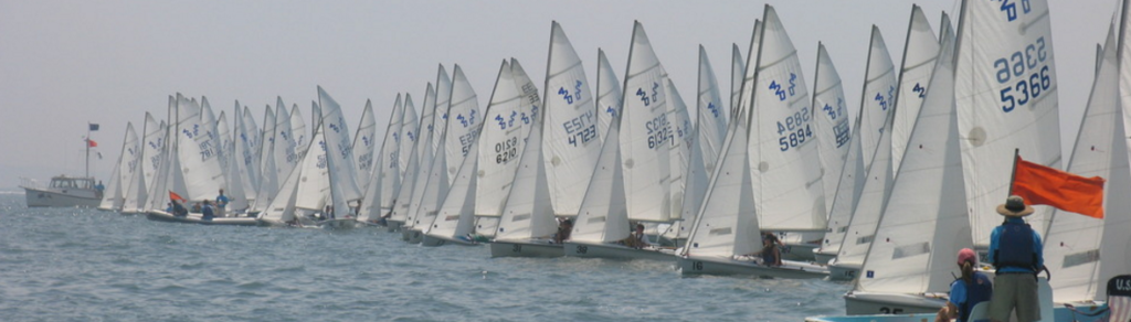 Dozens of small sailboats with white sails line up closely at the starting line of a regatta, with officials in boats nearby and flags signaling the race start on a calm, hazy day.