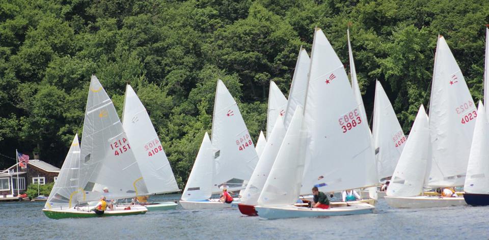 A group of small sailboats with white sails race closely together on a calm lake, with green trees and a building visible in the background. People are visible in each boat, focusing on steering.