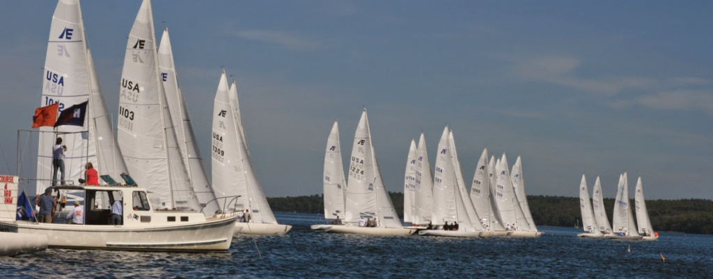 A fleet of white sailboats with numbered sails races on a lake under a blue sky, with a motorboat and people observing at the left side of the image. Trees line the distant shoreline.