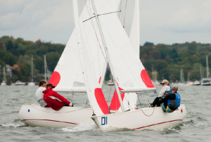 Two small sailboats with white sails and red markings are racing closely on the water, each with a crew of two. There are trees and anchored boats in the background under a cloudy sky.