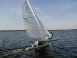 A person sails a small white catamaran with a single sail over calm water, leaning back as the boat glides quickly, creating a small wake. The sky is clear and the water stretches into the distance.